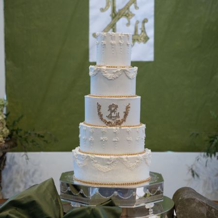 A white five tiered wedding cake with unique fancy piping and gold monogram with gold laurel leaf